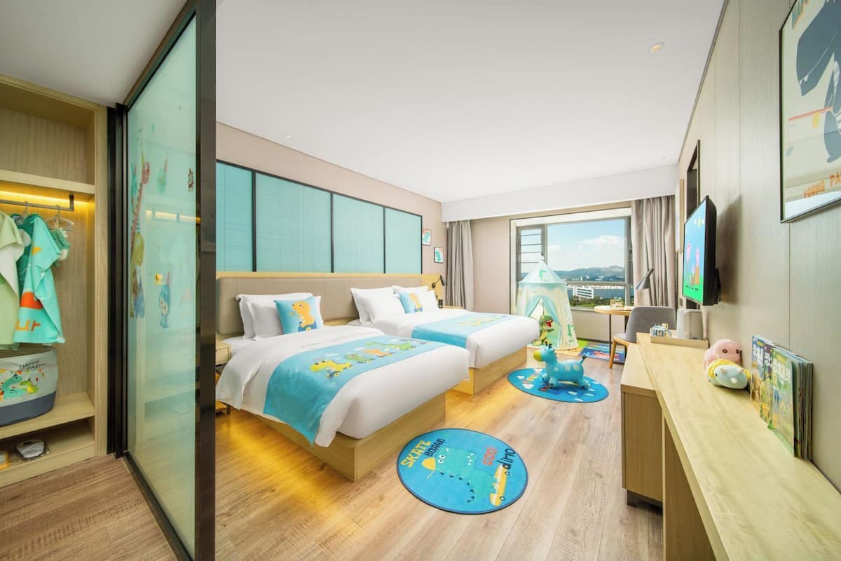 A hotel room with two beds, a desk, a chair, a TV, and a window with a view.