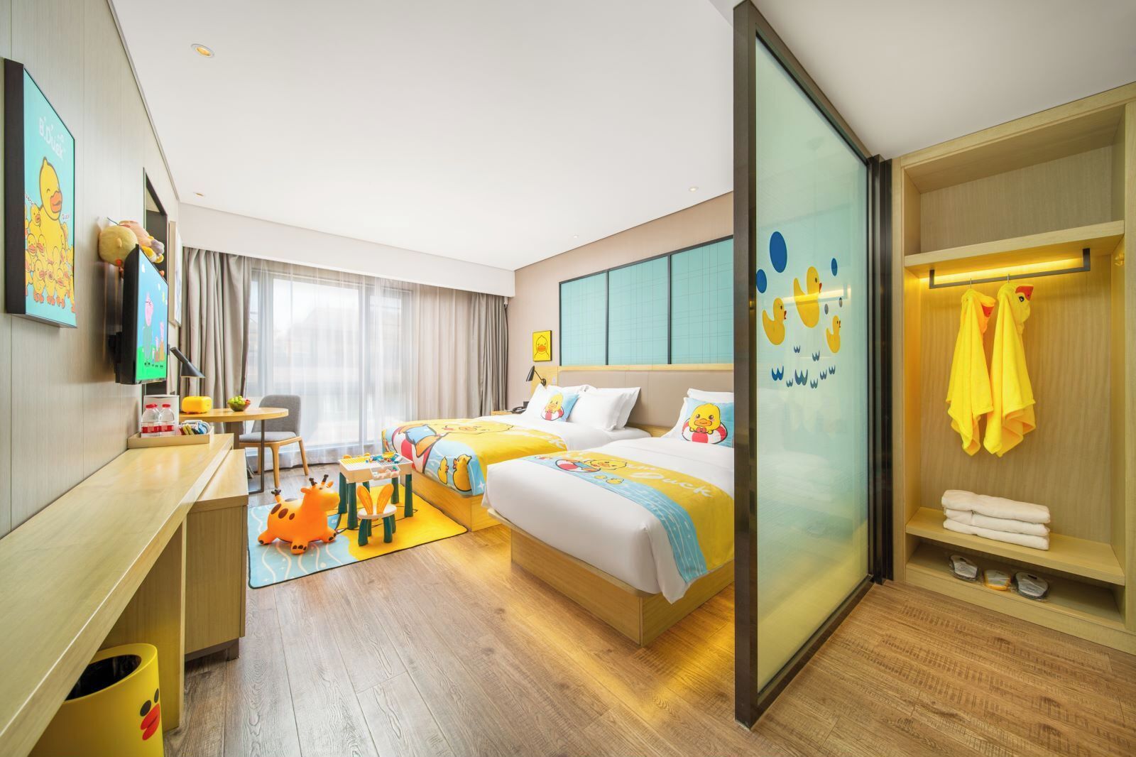 A hotel room with two beds, a desk, a chair, a small table, a sofa, a TV, a window with curtains, and a bathroom with a towel rack.