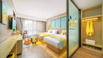A hotel room with two beds, a desk, a chair, a small table, a sofa, a TV, a window with curtains, and a bathroom with a towel rack.