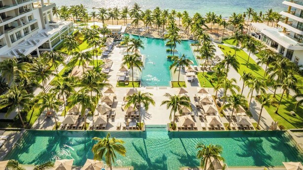 An aerial view of a resort with a central swimming pool surrounded by lounge areas and palm trees, overlooking a beach and ocean. - Confirmed Reservation in Cancun!Thanksgiving Week!   (Cancún)