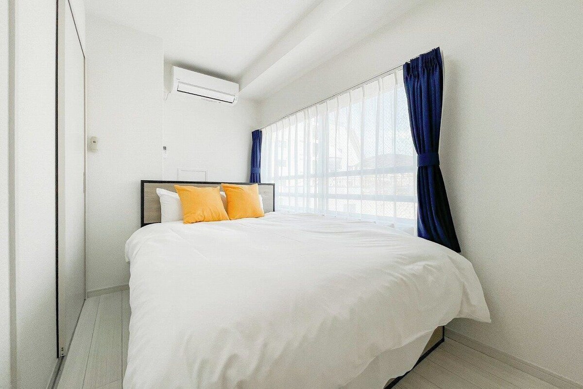 A bedroom with a bed, white walls, a window with blue curtains, and an air conditioning unit.