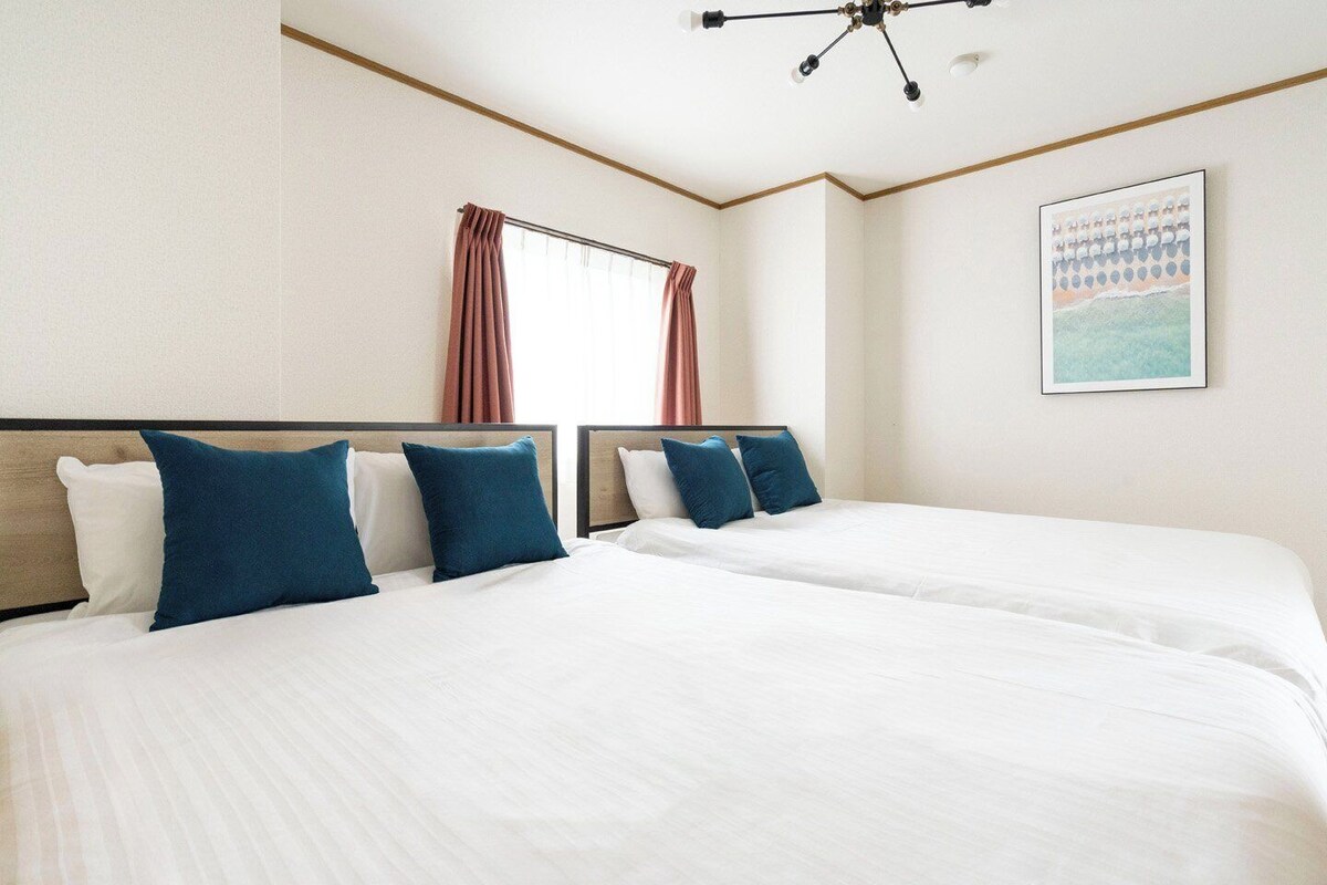 A hotel room with two beds, a large window with curtains, a ceiling light fixture, and a framed picture on the wall.
