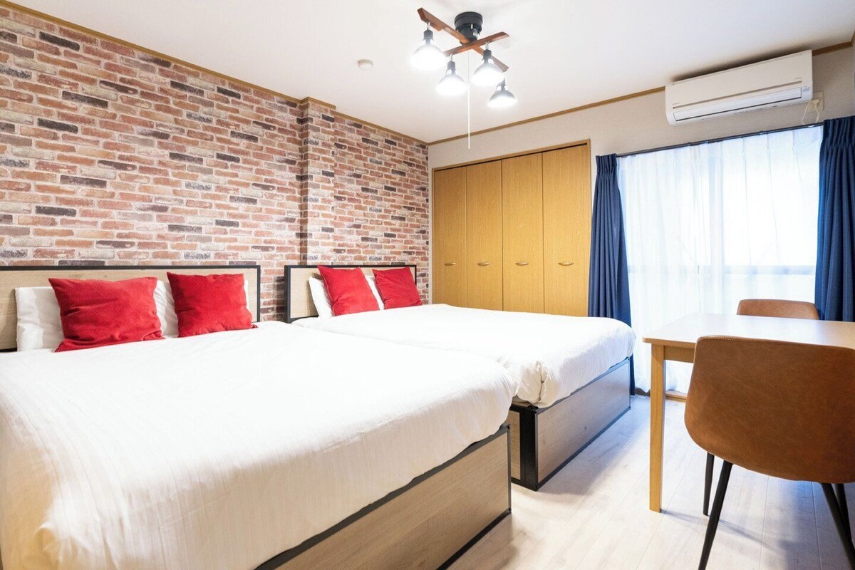 A hotel room with a brick wall, a large bed with red pillows, a wooden wardrobe, a desk with a chair, and an air conditioning unit.