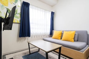 Ein Zimmer mit einem grauen Sofa, zwei gelben Kissen, einem kleinen Holztisch, einem an der Wand montierten Fernseher und einem Fenster mit blauen Vorhängen.