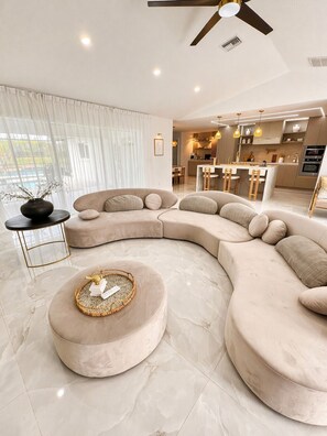 Ein modernes Wohnzimmer mit einer gebogenen Eckcouch, einem runden Hocker und einem Essbereich im Hintergrund.
