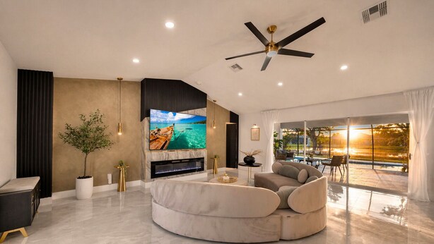 A modern living room with a large TV, a ceiling fan, and a view of a pool. - Luxury Canal Villa, Headed Pool, Game Room, Sandy Beach Yard & Fire Pit (Cape Coral)