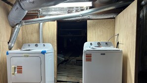 Deux machines à laver Maytag blanches se trouvent dans une buanderie aux murs en bois, aux conduits métalliques et au plancher en bois.