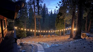 Une cabane en bois dotée d’une terrasse, entourée d’une forêt. On y trouve des guirlandes lumineuses, du mobilier d’extérieur et un chemin pavé de pierres.
