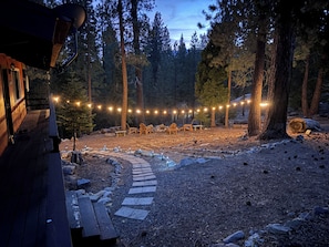 A wooden cabin with a deck, surrounded by a forest. There are string lights, outdoor furniture, and a stone pathway.