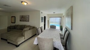 Una sala de estar con un sofá modular, una mesa de comedor con sillas y vistas a una piscina a través de un ventanal.