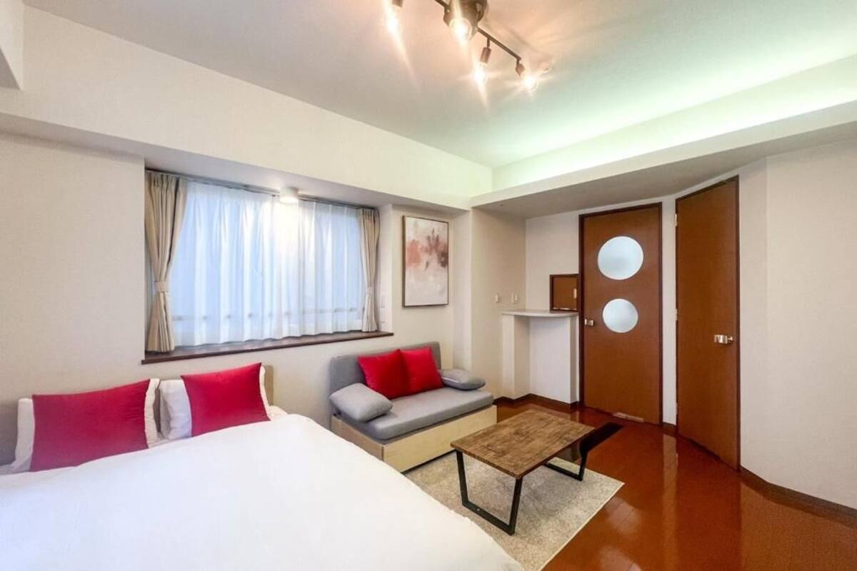 A hotel room with a bed, a sofa, a small table, a window with curtains, a door with a round mirror, and a wall-mounted light fixture.