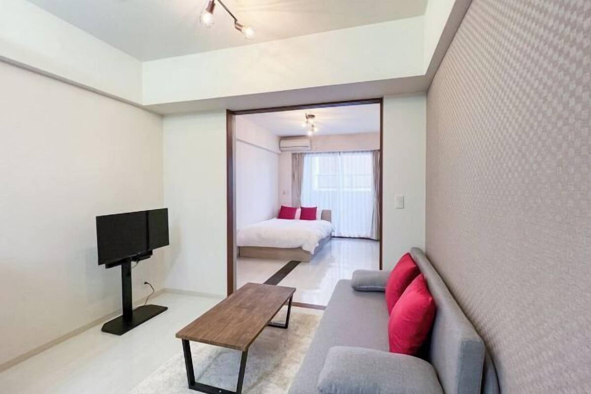 A modern hotel room with a flat-screen TV, a sofa with red cushions, a wooden coffee table, and a view into the adjacent bedroom.