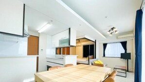 A compact room with a wooden dining table, chairs, a kitchenette, a small seating area, and a large window with curtains. - CITYSPIRE Sakurakawa II (Osaka)