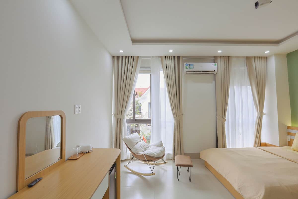 Habitación de hotel con cama, sillón mecedora, una mesita con control remoto y ventana con cortinas.