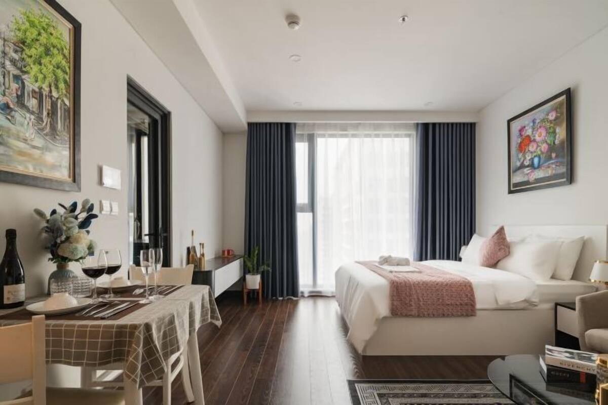 A modern hotel room with a bed, bedside table, and a dining area with a table set for two.