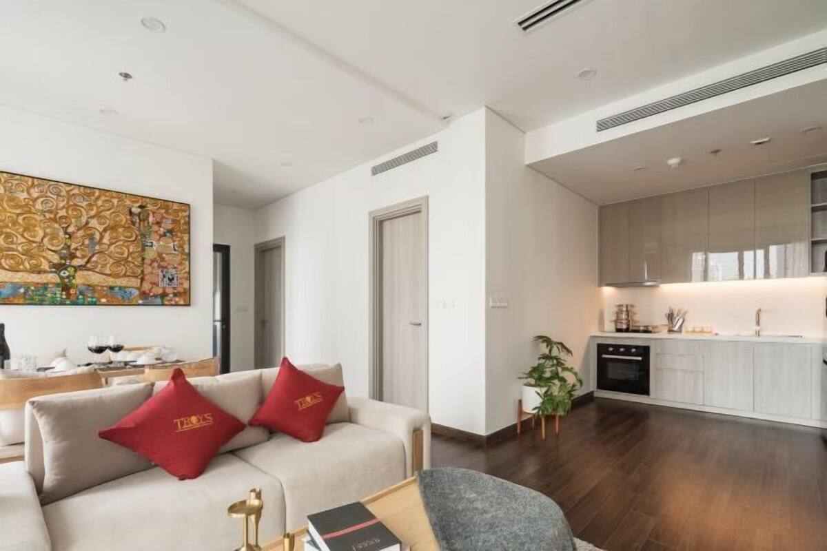 A modern living room with a sofa, a coffee table, and a dining area with a table and chairs. There is a kitchen with white cabinets and a stove.
