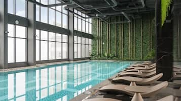 An indoor swimming pool with clear blue water, surrounded by large windows and a wall of green foliage.