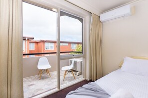 A hotel room with a bed, a window with curtains, a small table with a chair, and an air conditioning unit. - Retro 3BR Mid Century Apartment (St Kilda East)