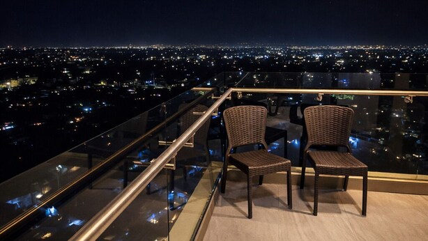 Uma varanda com cadeiras de vime, guarda-corpo de vidro e vista noturna da cidade.