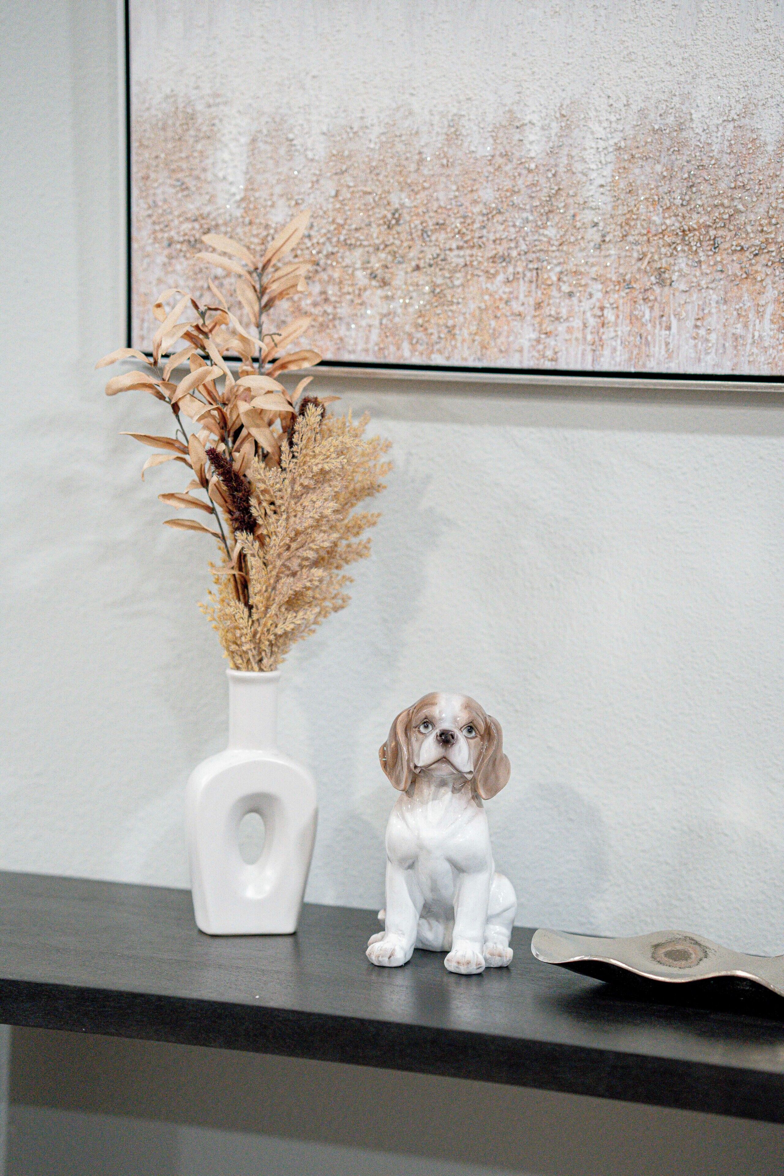 Une statuette de chien en céramique blanche, un vase blanc contenant des plantes séchées et une œuvre d’art encadrée, le tout posé sur une étagère.