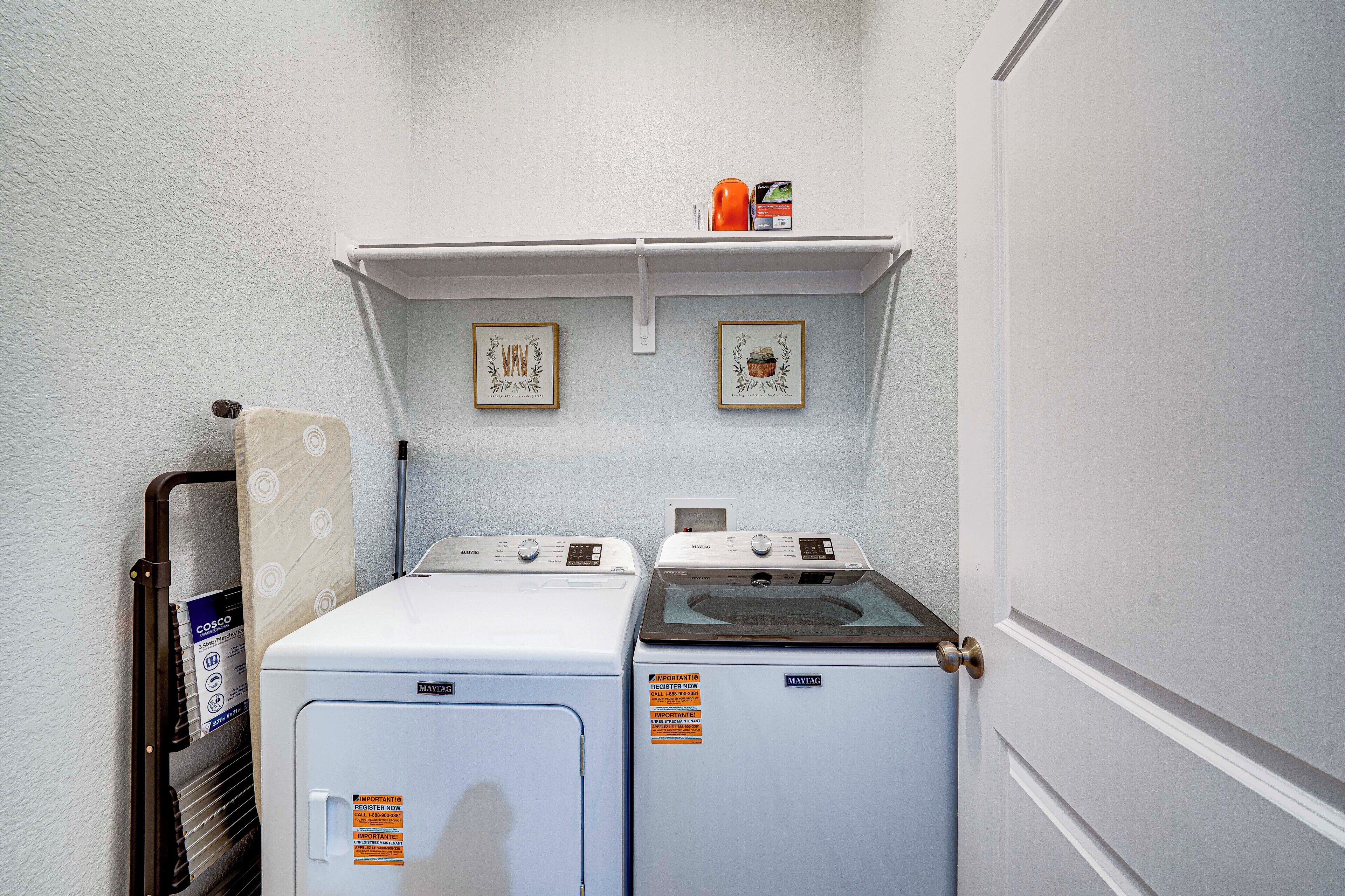 Une buanderie équipée de deux machines à laver et deux sèche-linge, une étagère avec divers objets et des tableaux encadrés accrochés au mur.