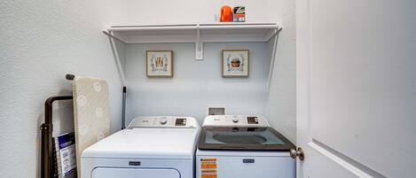 Une buanderie équipée de deux machines à laver et deux sèche-linge, une étagère avec divers objets et des tableaux encadrés accrochés au mur.