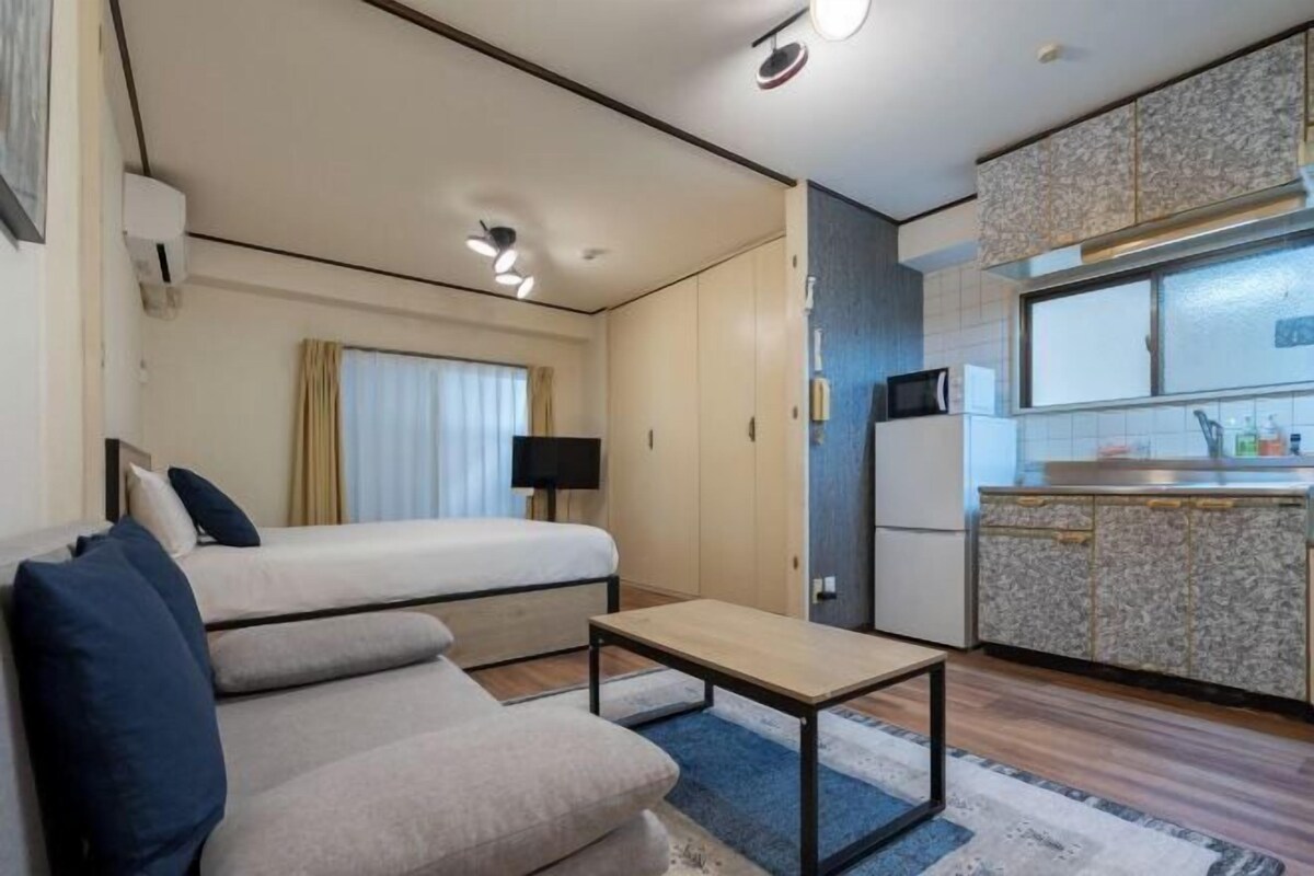 A compact hotel room with a bed, sofa, small table, kitchenette, and a window with curtains.