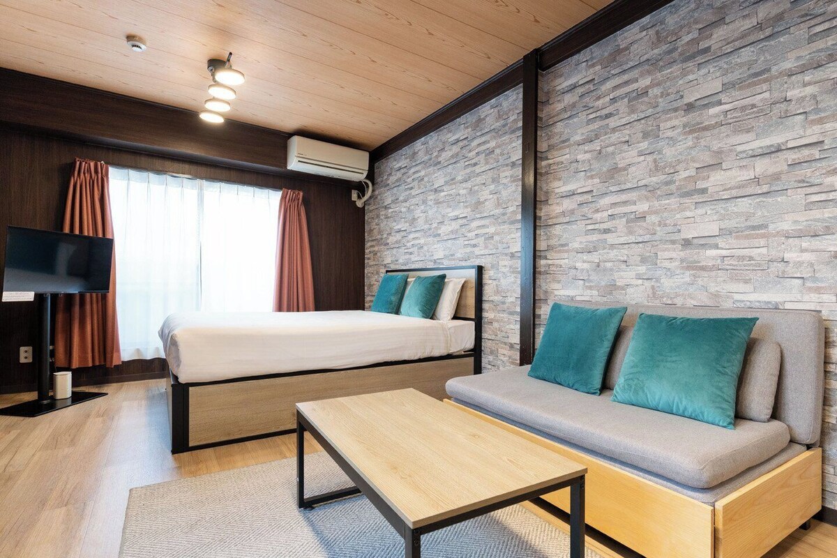 A modern hotel room with a bed, sofa, TV, and stone wall.