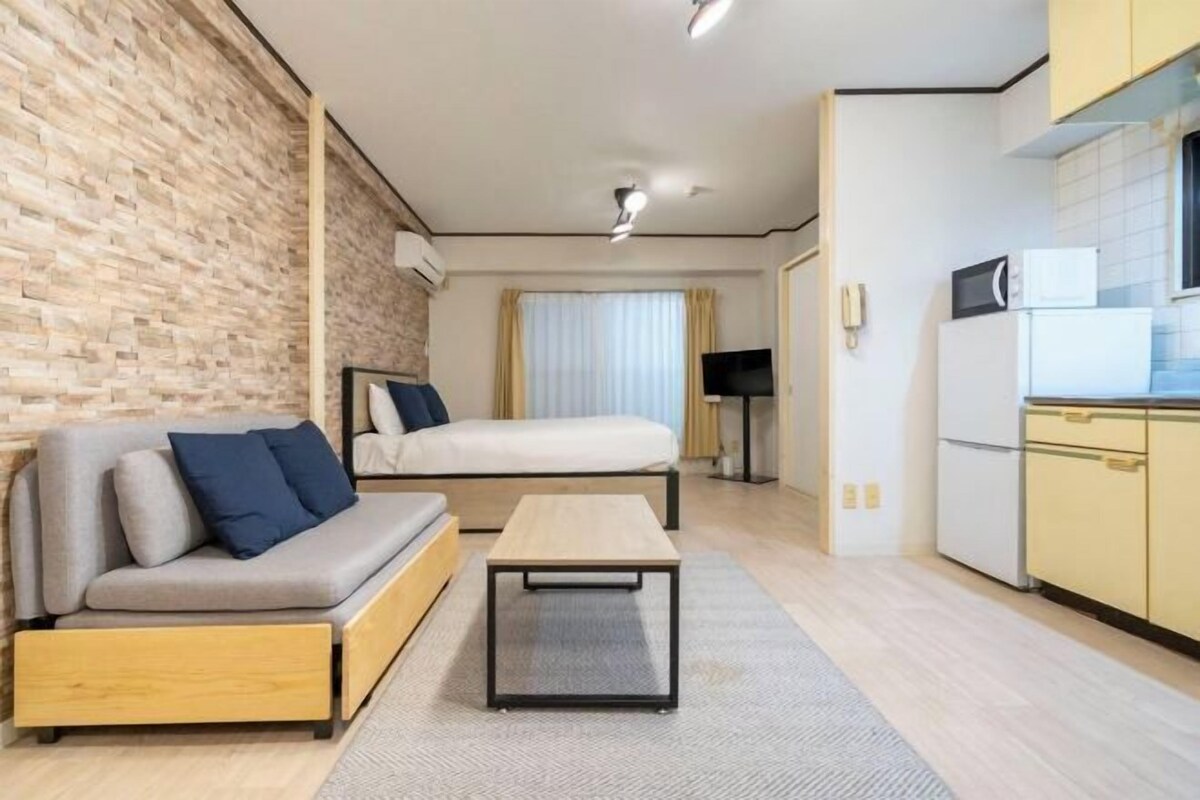 A compact living space with a sofa, a small table, a bed, a TV, and a kitchen area with a microwave.