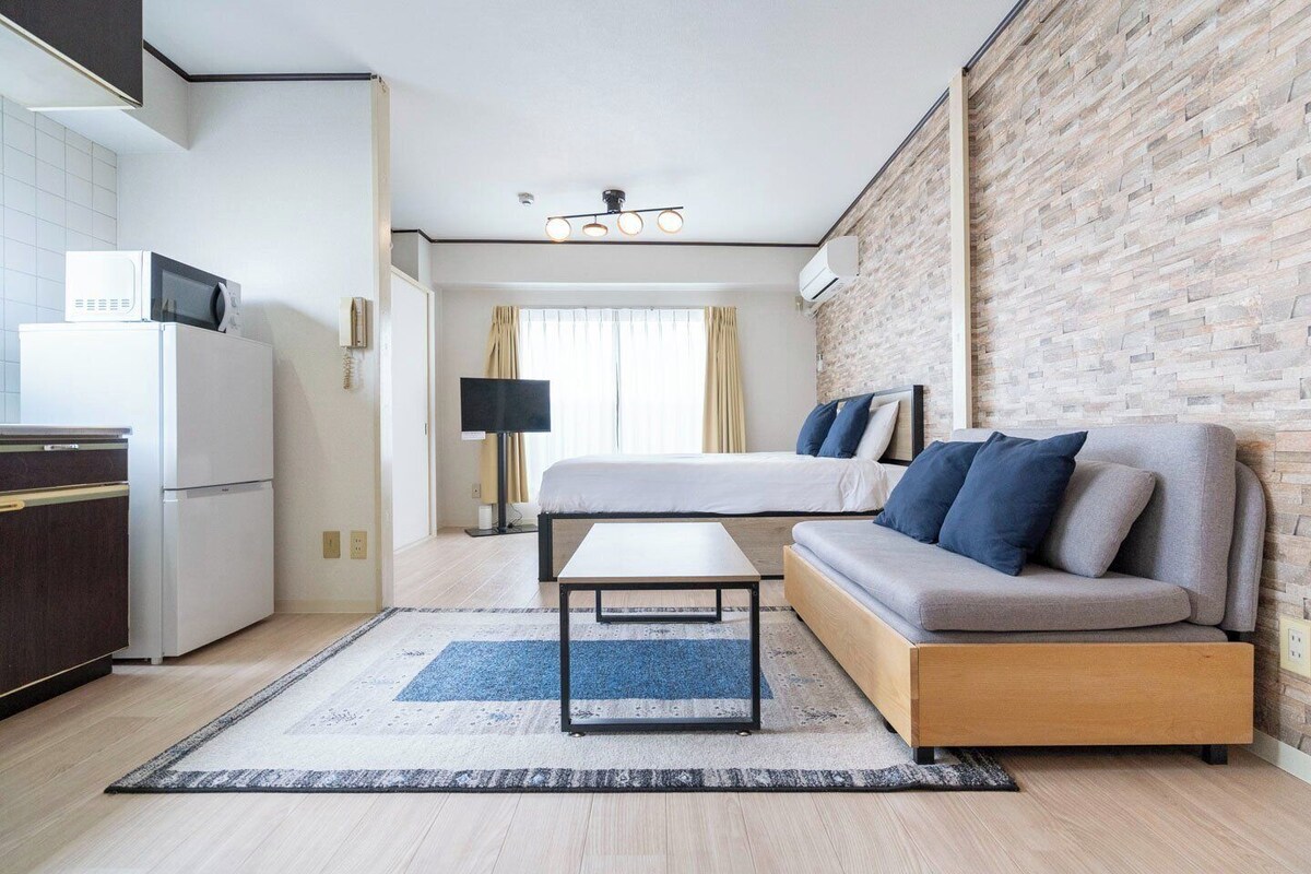 A modern hotel room with a kitchenette, a bed with blue and grey bedding, a wooden sofa, and a brick accent wall.