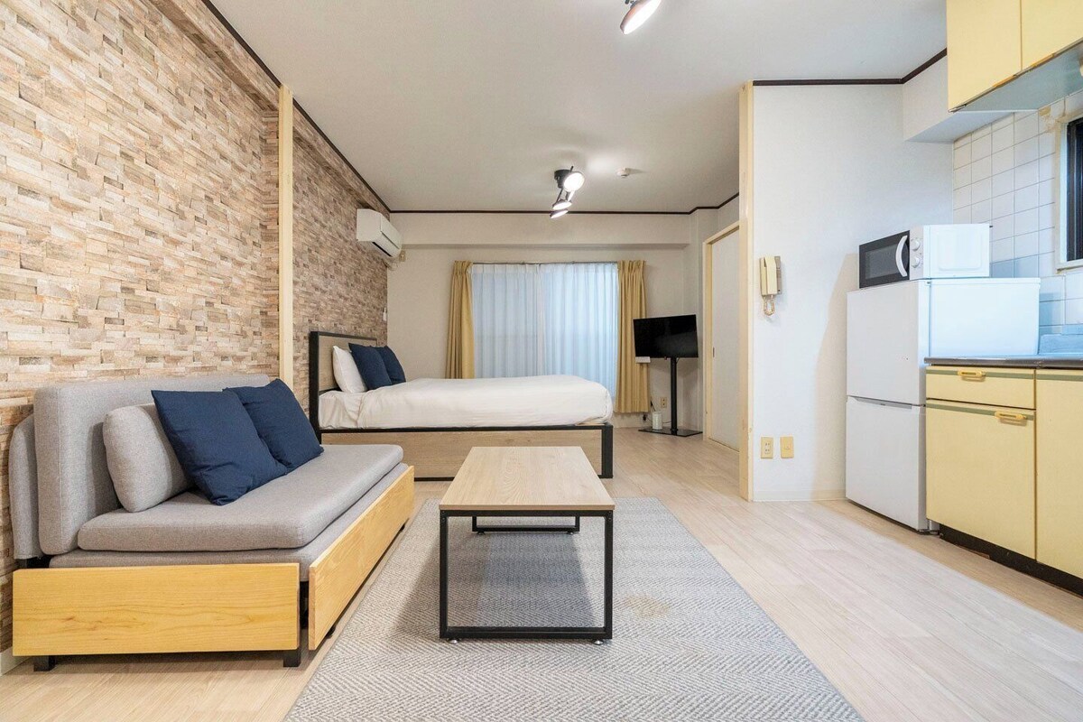 A compact room with a sofa, a bed, a small table, a TV, and a kitchenette.