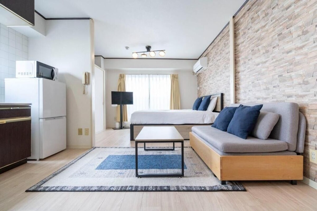 A modern hotel room with a kitchenette, a bed with a headboard, a sofa, a flat-screen TV, and a large window.