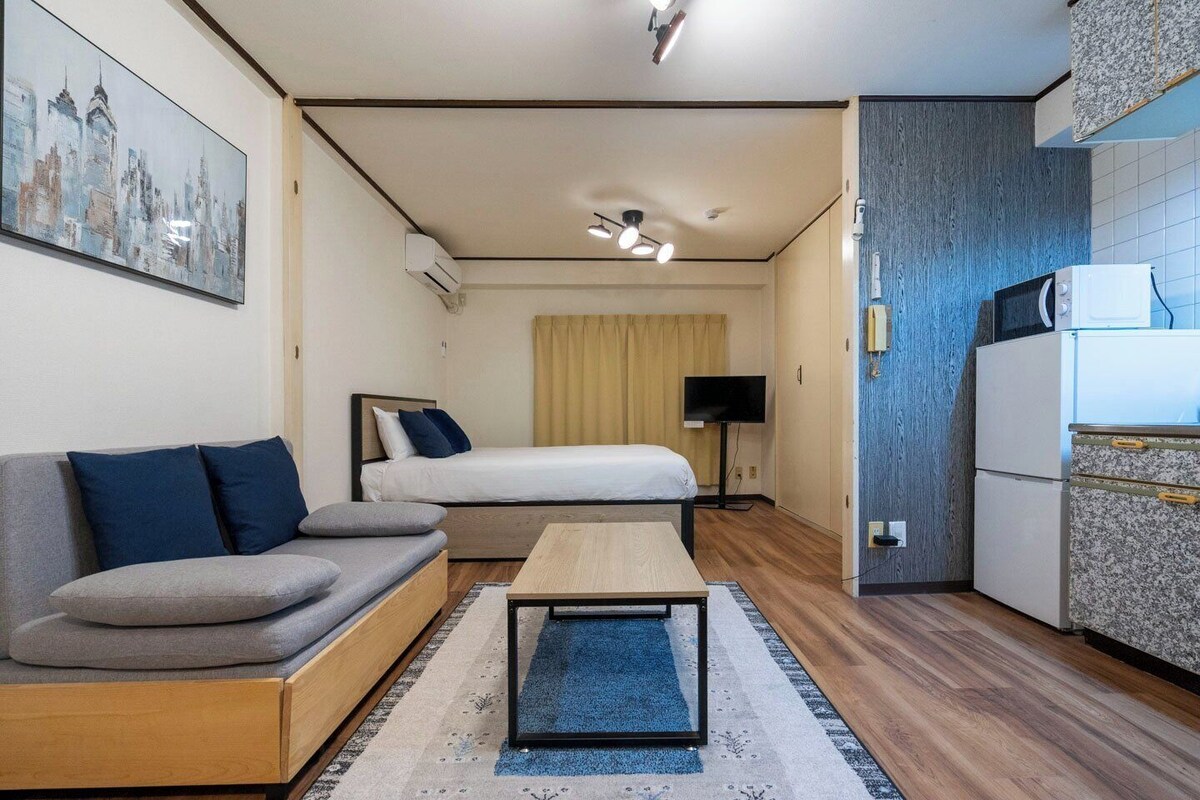 A compact hotel room with a sofa, a bed, a small table, a television, and a kitchenette.