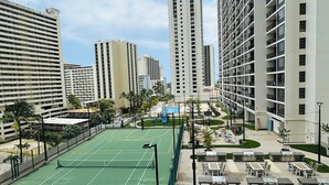 Una cancha de tenis rodeada de edificios altos, mobiliario exterior y una zona de piscina.