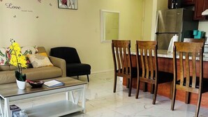Sala de estar con sofá, mesa de centro y sillas, cocina con refrigerador y armarios, y pared decorada.