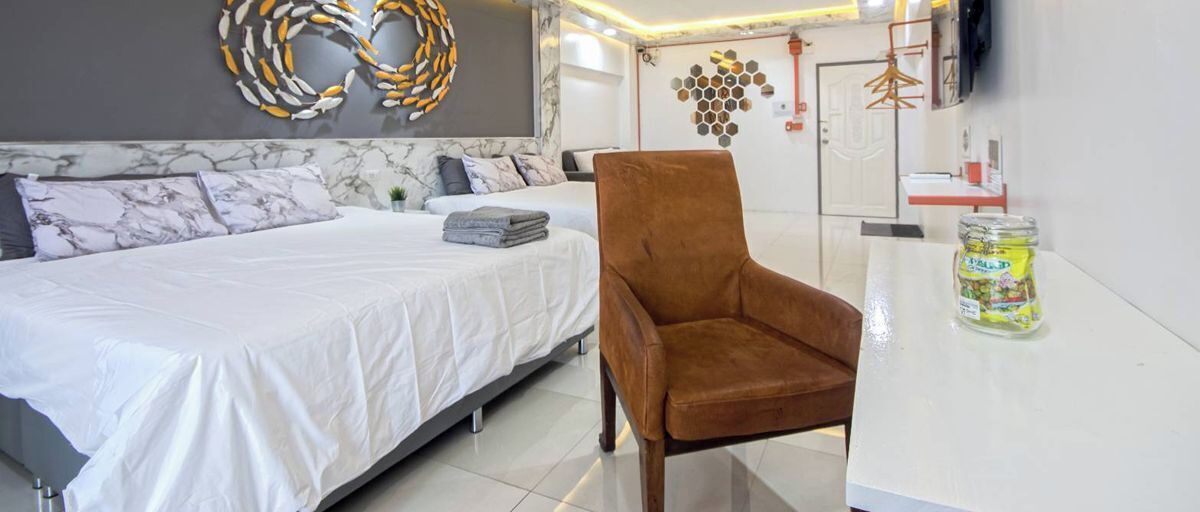 A hotel room with a bed, a chair, a TV, a wall art piece, and a small table with a jar.