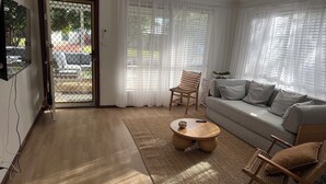 A living room with a sofa, a coffee table, a wicker chair, and a large window with sheer curtains. - Villa Quindy - ideally located a short 2 minute stroll to Quindalup bay beach. (Quindalup)