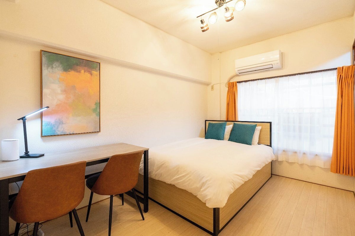 A hotel room with a bed, desk, chairs, an air conditioner, and a painting on the wall.