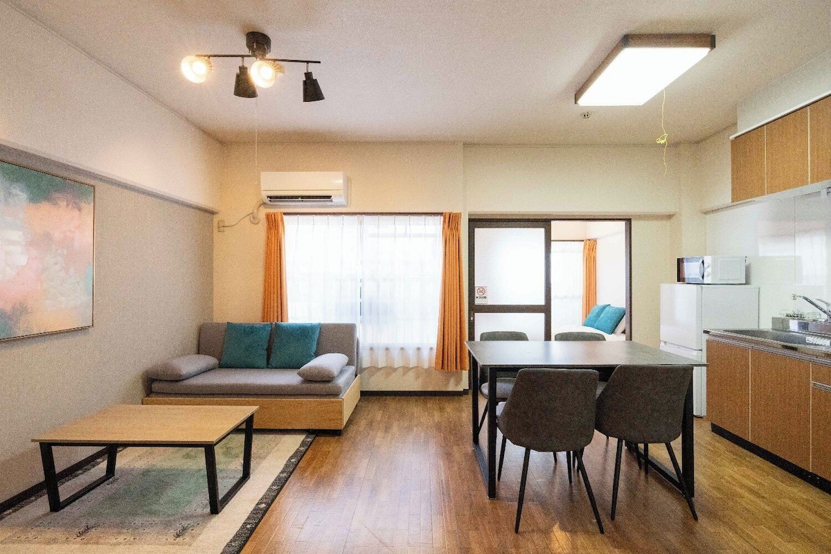 A compact living space with a sofa, dining table, chairs, a small kitchen area, and an air conditioning unit.