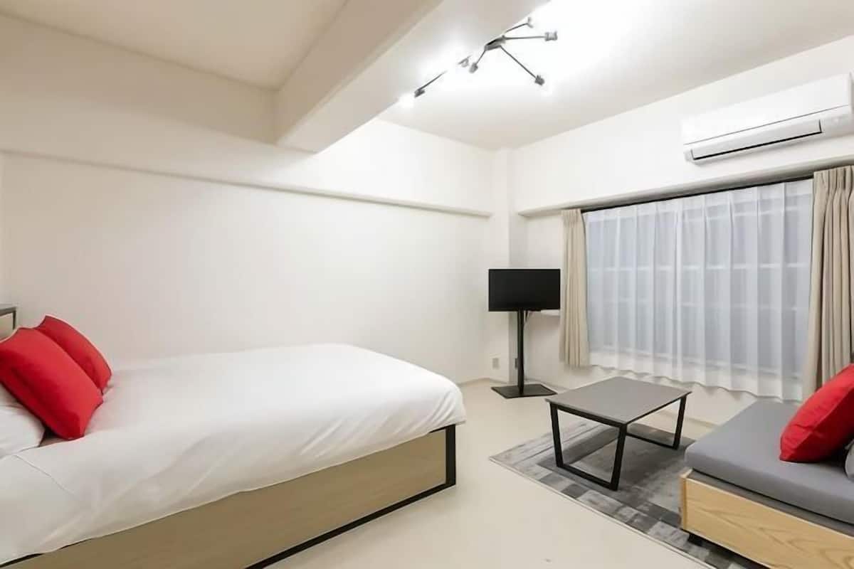 A modern hotel room with a bed, a small table, a sofa, a TV, and an air conditioning unit.