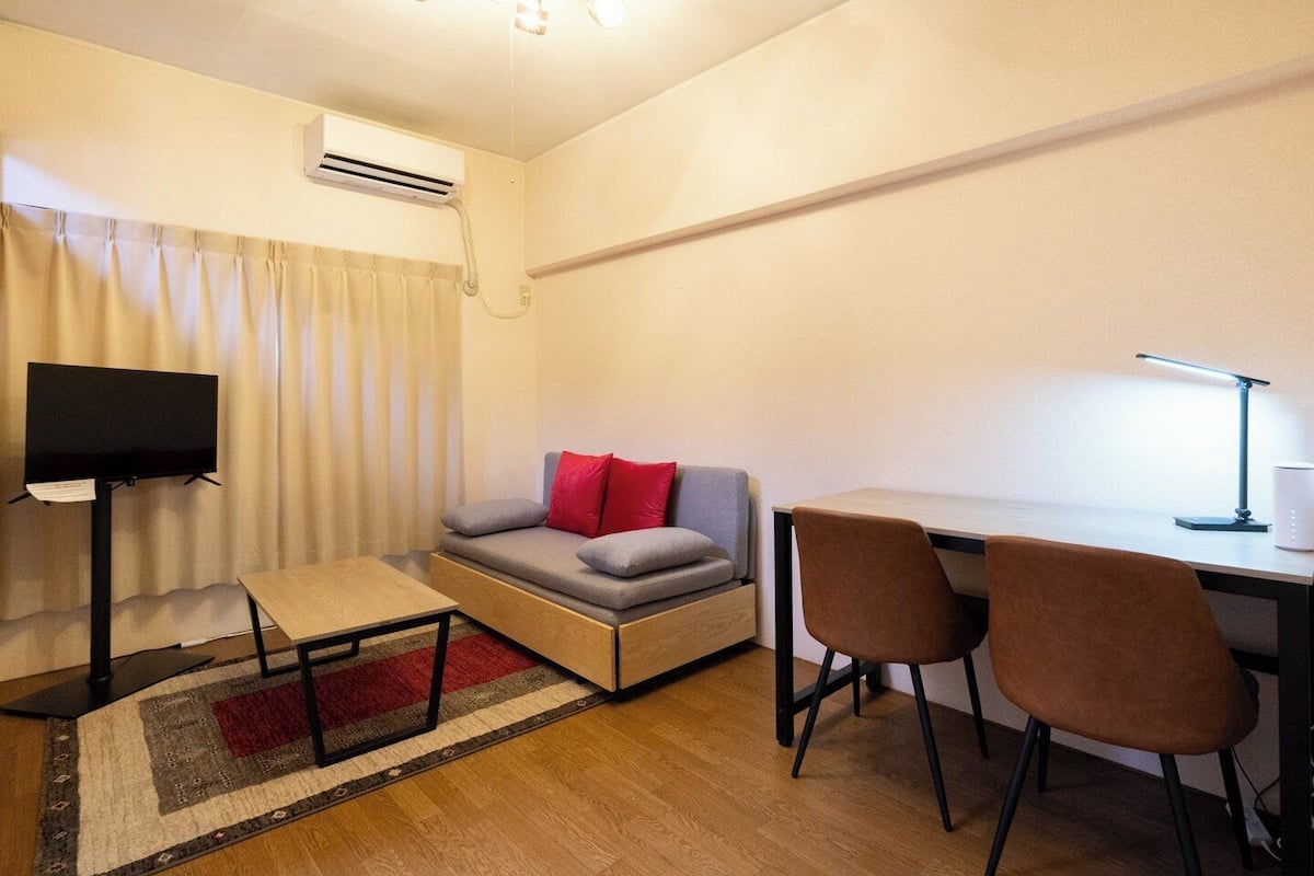 A compact room with a sofa, a small table, a desk with two chairs, a TV on a stand, and an air conditioning unit.