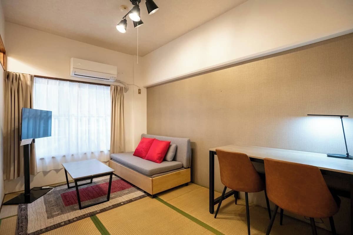 A compact room with a sofa, a desk, two chairs, a standing lamp, and an air conditioning unit.