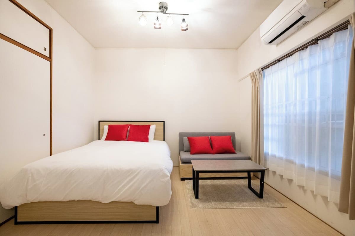 A bedroom with a bed, a sofa, a coffee table, an air conditioning unit, and a window with curtains.