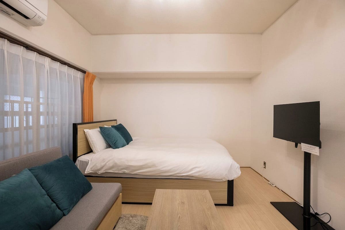 A small, modern room with a bed, a sofa, a TV, and a window with curtains.