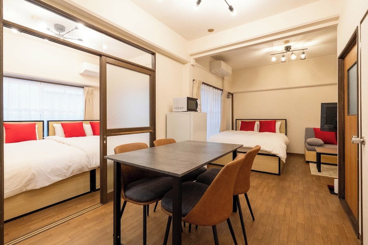 A hotel room with two beds, a dining table, chairs, a TV, and an air conditioning unit.