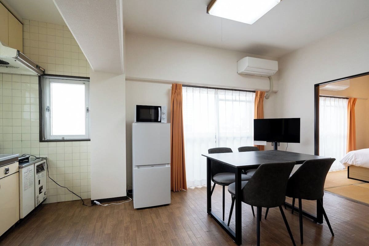A compact hotel room with a kitchenette, a dining table with chairs, a flat-screen TV, and a bed.