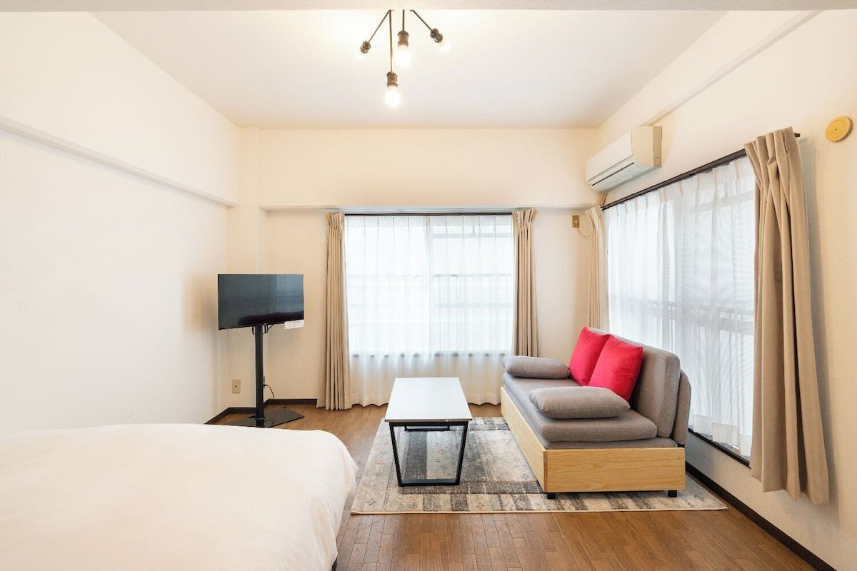 A hotel room with a bed, a sofa with red cushions, a coffee table, a TV, and large windows with curtains.