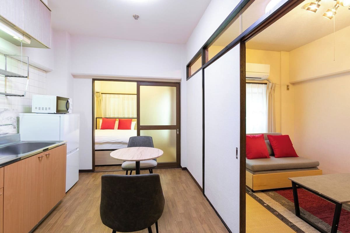 A compact hotel room with a kitchenette, a bed with red pillows, a small round table, and a sofa with red cushions.