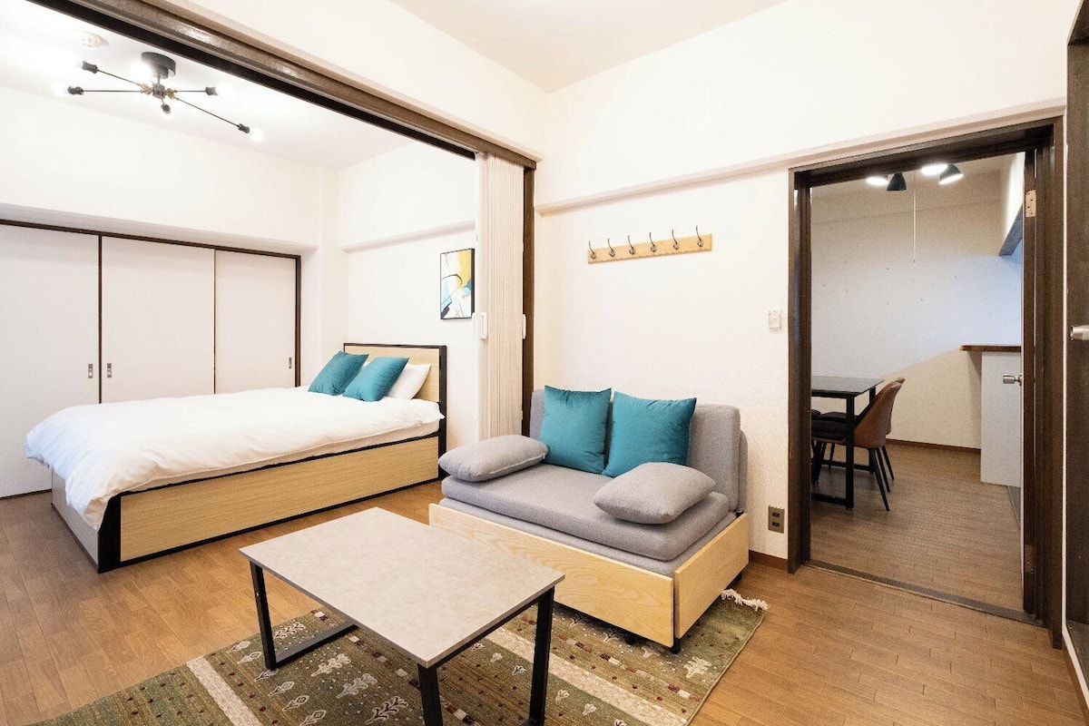 A compact room with a bed, a sofa, a small table, and a kitchenette area.