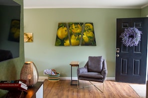 Ein Wohnzimmer mit einer dunkelbraunen Tür, einem grauen Sessel, einem kleinen Tisch, einem an der Wand hängenden Bild mit Zitronen und einem dekorativen Kranz.